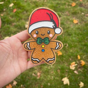 Puede incluir: Adorno navideño de hombre de jengibre con gorro de Papá Noel rojo, pajarita verde y una cara sonriente. El adorno es marrón con contornos negros y un aspecto texturizado. El fondo es hierba verde con hojas caídas.