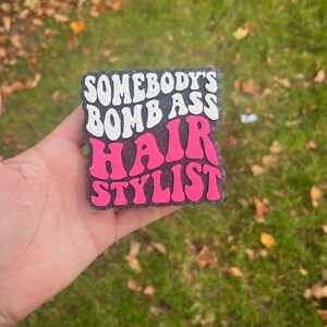 Op de afbeelding: Een rechthoekige, zwart-paarse luchtverfrisser met de tekst "SOMEBODY'S BOMB ASS HAIR STYLIST" in witte en roze bubbelletters. De luchtverfrisser wordt in een hand gehouden tegen een achtergrond van groen gras en gevallen bladeren.