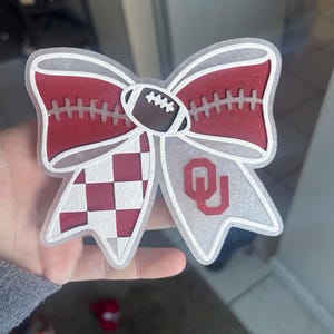May include: A decorative bow featuring a football and the OU logo. The bow has a red and white colour scheme, with a football in the centre and a chequered pattern on one side. The other side displays the OU logo.
