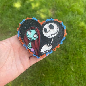 Op de afbeelding: Een hartvormige decoratie met Sally en Jack Skellington uit The Nightmare Before Christmas. De personages staan tegen een zwarte achtergrond met een oranje en blauwe rand. De decoratie wordt tegen een achtergrond van groen gras gehouden.