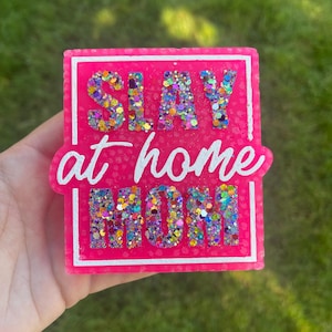 Puede incluir: Una pieza rectangular de resina rosa intenso con las palabras "SLAY at home MOM" en escritura blanca. Las letras están rellenas de purpurina colorida. La pieza se sostiene sobre un fondo verde borroso.