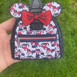 May include: A black and white Minnie Mouse-themed backpack with a red glitter bow and Minnie Mouse ears. The backpack features a pattern of Minnie Mouse on a white background with red and navy blue accents. It has a zippered front pocket.