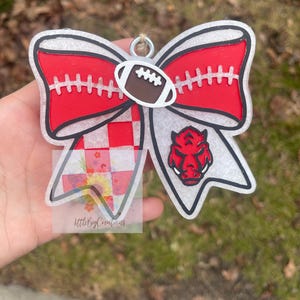 May include: A clear, bow-shaped ornament with a red and white design. The bow features a football in the centre and a red Razorback logo on the right. The left side has a red and white checkered pattern. The text "littleBigCreations" is visible.