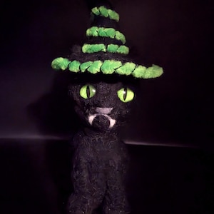 May include: A black cat figurine wearing a witch's hat with green trim. The cat has glowing green eyes and a small white patch on its face. The figurine is set against a dark background, creating a spooky Halloween theme.