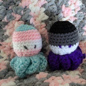 May include: Two crocheted octopus plushies. The first octopus is light blue with a pink, white, and light blue striped head. The second octopus is purple with a black, gray, and white striped head.