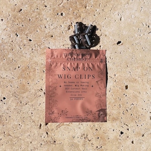 May include: A package of snap-on wig clips from The Smart Chemist. The pink package has the words "SNAP ON WIG CLIPS" printed on it. The package also states that the clips are for wig making and converting hair extensions. There are 16 pieces in the package.