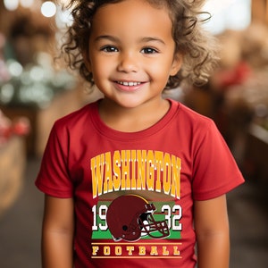 May include: A child wearing a maroon t-shirt with a football design. The shirt displays the text "WASHINGTON 1932 FOOTBALL" in gold and white, and a maroon helmet. The back print is available.