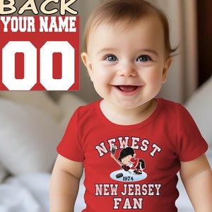 Op de afbeelding: Een rode baby romper met een cartoon hockey speler en de tekst "NEWEST 1974 NEW JERSEY FAN". De romper heeft een wit nummer "00" op de rug met de tekst "BACK YOUR NAME".