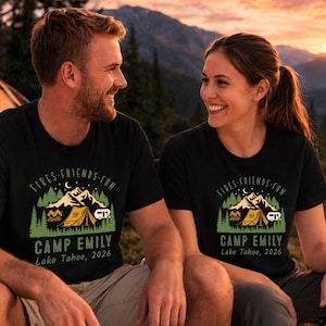 Könnte beinhalten: Ein schwarzes Kurzarm-T-Shirt mit einem Grafikdruck einer Berglandschaft, einem Zelt, einem Wohnwagen und Tannenbäumen. Der Text "FIRES-FRIENDS-FUN", "CAMP EMILY" und "Lake Tahoe, 2026" ist in grüner und weißer Schrift auf der Vorderseite zu sehen.