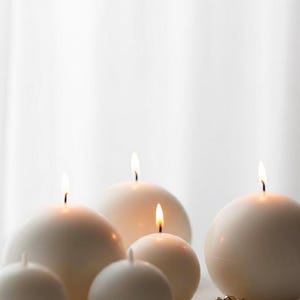 May include: A collection of white, spherical candles of varying sizes, some lit with small flames. The candles are arranged in a group, with a few small gold bells in the foreground. The background is a soft, white, out-of-focus area.