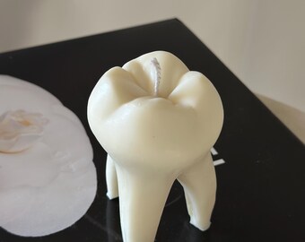 Pode incluir: Uma vela branca em forma de dente com um pavio branco. A vela está sobre uma superfície preta.