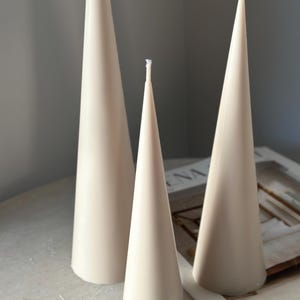May include: Three beige cone-shaped candles of varying sizes. The candles have a smooth, matte finish. The wicks are visible at the top. The candles are arranged on a light-coloured surface.