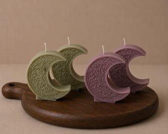 May include: Four crescent moon-shaped candles featuring intricate embossed mandala patterns. Two candles are sage green and two are dusty mauve, each topped with a single white wick. The candles are arranged in pairs on a dark wooden board.