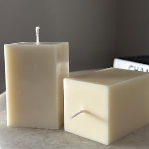 Puede incluir: Dos velas de pilar cuadradas de color marfil. Una vela está en posición vertical y la otra en horizontal. Ambas tienen mechas blancas. Las velas están sobre una superficie clara, posiblemente de mármol. El fondo es gris neutro.