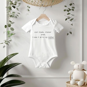 May include: White baby onesie with short sleeves, hanging on a wooden hanger. The onesie has the text "cool shade, stunner yeah, I owe it all to my mother" printed in black. The background includes greenery and a stuffed animal.
