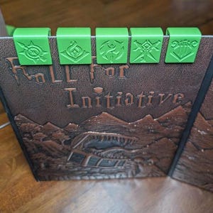 May include: A brown leather-bound game book with the words "Roll for Initiative" embossed on the cover. Five green plastic clips with various symbols are attached to the top edge of the book. The book has a detailed landscape design.