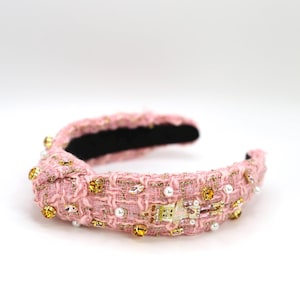 May include: Pink tweed headband with gold and iridescent embellishments. The knotted headband features a black interior and is adorned with faux pearls, crystals, and gold-toned accents. A stylish hair accessory.