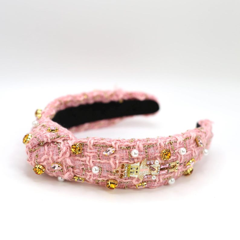 May include: Pink tweed headband with gold and iridescent embellishments. The knotted headband features a black interior and is adorned with faux pearls, crystals, and gold-toned accents. A stylish hair accessory.