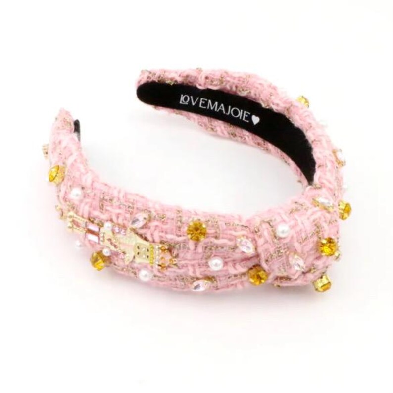May include: Pink tweed headband with gold and clear crystal embellishments, pearls, and a knotted design. The headband features the text "LOVE MAJOIE" on the inner band. A stylish hair accessory.
