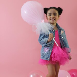 May include: A denim jacket with a sequined heart patch, a bright pink leotard, and a layered tulle skirt. The outfit is paired with metallic gold boots and a large, light pink tulle-covered balloon on a white stick, creating a festive look.