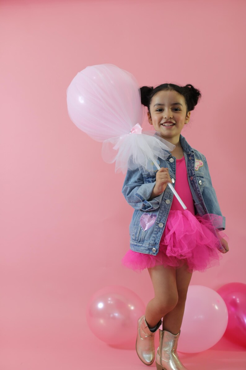 May include: A denim jacket with a sequined heart patch, a bright pink leotard, and a layered tulle skirt. The outfit is paired with metallic gold boots and a large, light pink tulle-covered balloon on a white stick, creating a festive look.