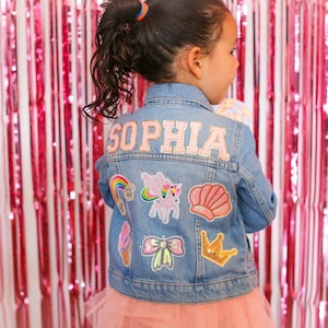 May include: A blue denim jacket with the name "Sophia" embroidered on the back. The jacket is decorated with various colorful patches, including a rainbow, a unicorn, a seashell, a bow, a crown, and an ice cream cone.