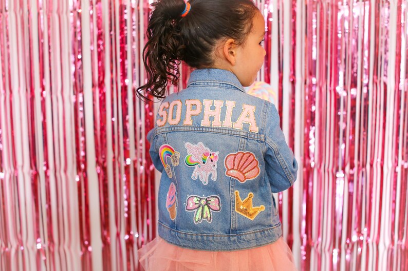 May include: A blue denim jacket with the name "Sophia" embroidered on the back. The jacket is decorated with various colorful patches, including a rainbow, a unicorn, a seashell, a bow, a crown, and an ice cream cone.
