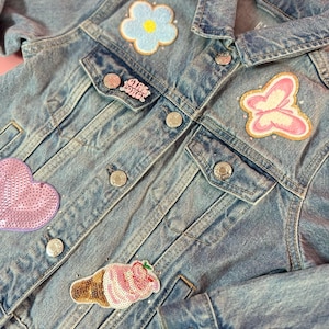 May include: A light blue denim jacket with various patches, including a flower, a butterfly, a heart, an ice cream cone, and a pink pin that says "Girl Power".