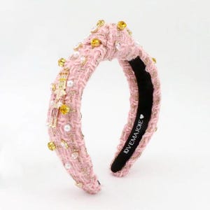 May include: Pink tweed headband adorned with gold-toned embellishments, including crystals and pearls. The headband features a padded design for comfort and a black interior with the text "OVEMAJOIE". A stylish hair accessory.