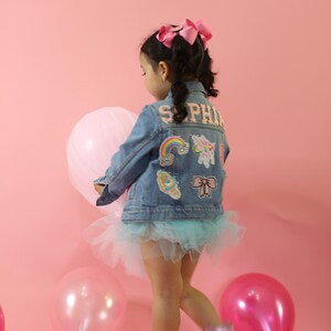 May include: A blue denim jacket featuring the name SOPHIA in block letters, decorated with colorful patches including a rainbow, unicorn, ice cream, and bow. The jacket is worn over a light blue tulle skirt with pink glittery boots and a pink hair bow.