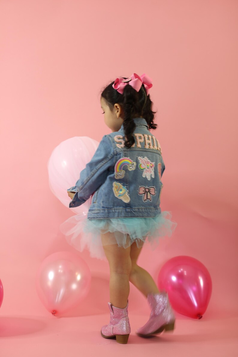 May include: A blue denim jacket featuring the name SOPHIA in block letters, decorated with colorful patches including a rainbow, unicorn, ice cream, and bow. The jacket is worn over a light blue tulle skirt with pink glittery boots and a pink hair bow.