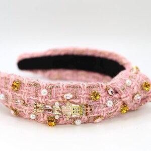 May include: Pink tweed headband with gold accents, pearls, and jewel embellishments. The headband features a decorative knot and a small, detailed charm. The interior is lined with black fabric, creating a stylish accessory.