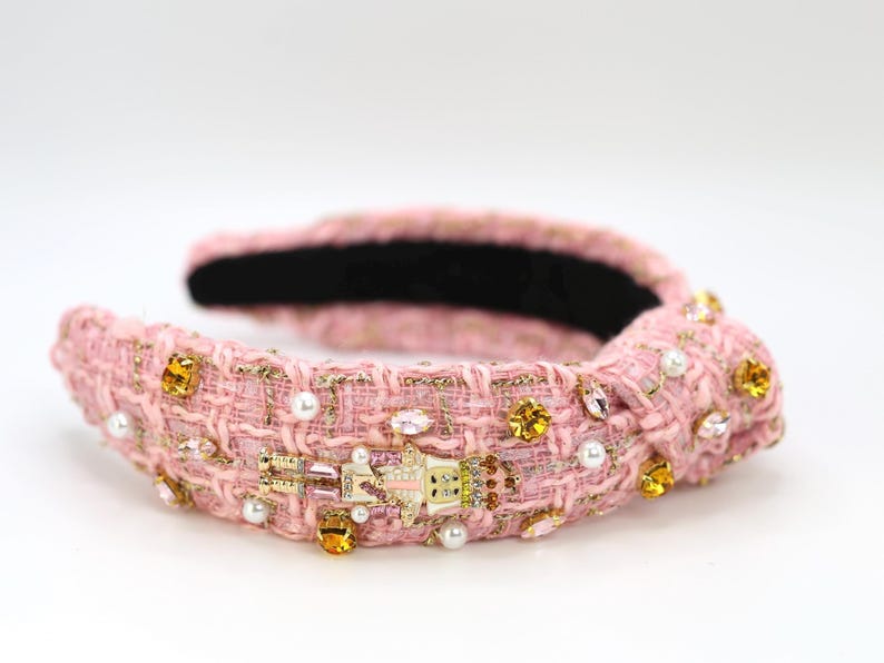 May include: Pink tweed headband with gold accents, pearls, and jewel embellishments. The headband features a decorative knot and a small, detailed charm. The interior is lined with black fabric, creating a stylish accessory.