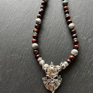 Op de afbeelding: Een ketting met een zilverkleurige Viking schedel hanger. De ketting is samengesteld uit afwisselend donkerbruine en grijze stenen kralen. De hanger heeft een gedetailleerd schedelontwerp met een gehoornde helm en een lange baard.