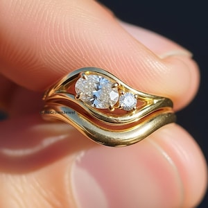 Könnte beinhalten: Ein goldener Verlobungsring-Satz mit wellenförmigem Design. Der Verlobungsring verfügt über einen großen Diamanten im Ovalschliff und einen kleineren runden Diamanten. Das Band ist aus glattem, poliertem Gold. Das Set enthält ein passendes Goldband.