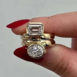 May include: Three gold rings with clear gemstones. The top ring features an emerald-cut stone, while the bottom ring has an oval-shaped stone. The rings are stacked together, showcasing their design and craftsmanship.