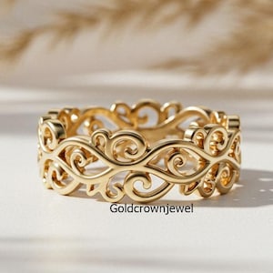 以下が含まれることがあります： 複雑な透かし彫りのデザインが施されたゴールドカラーのリング。リングは、渦巻くスクロールのような形状の繰り返し模様が特徴です。白い表面に、ニュートラルな背景で表示されています。リングはバンドスタイルです。