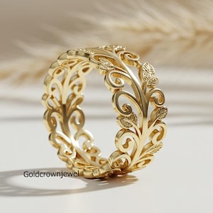 以下が含まれることがあります： 精巧なフローラルデザインのゴールドカラーのリング。リングは、葉と渦巻くツルの連続模様が特徴です。柔らかくニュートラルな背景の白い表面に表示されています。「Goldcrownjewel」の文字がリングに表示されています。