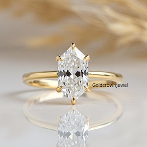 May include: A gold engagement ring featuring a large, clear, hexagonal-cut diamond. The ring has a thin, polished band. The diamond is the focal point, reflecting light. The background is soft and blurred.