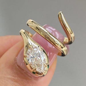 以下が含まれることがあります： 金色のヘビのデザインのリング。ヘビの頭として梨型の透明な宝石が特徴です。リングは滑らかで磨かれた仕上げで指に巻き付きます。小さな緑色の宝石がヘビの頭をアクセントにしています。