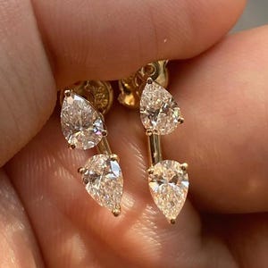 May include: A pair of gold earrings, each featuring two pear-cut clear gemstones arranged vertically on a thin metal bar. The stones are set in a prong setting, and the earrings have a polished gold finish, showcasing a delicate and refined design.