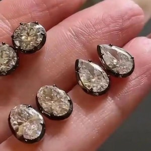 Könnte beinhalten: Mehrere diamantähnliche Edelsteine in ovaler und tropfenförmiger Form. Die klaren Steine sind in dunklen Metallfassungen gefasst. Die Edelsteine werden vor einem hellen Hintergrund präsentiert.