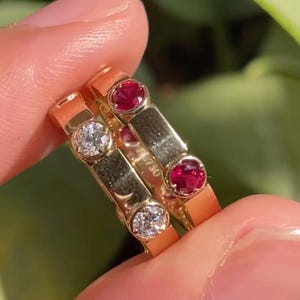 May include: Close-up of three gold rings. Two rings feature a single round, clear diamond, while the third ring has two round, red gemstones. The rings are held between fingers against a blurred green background.