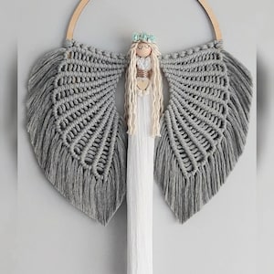 Op de afbeelding: Een handgemaakte macramé engel wanddecoratie. De engel heeft grijze vleugels, een houten hoofd met een bloemenkrans en lang, vloeiend wit haar. Het lichaam van de engel is een lang, wit, getextureerd stuk.