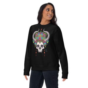 Puede incluir: Sudadera negra con un diseño detallado de calavera con cuernos, una corona floral y cuentas colgantes. La calavera tiene dientes afilados y está adornada con flores coloridas. El diseño está centrado en la parte delantera de la sudadera.