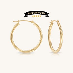 May include: A pair of polished gold hoop earrings with a smooth, rounded profile and a secure hinged clasp mechanism. Above the earrings, a black banner displays the text Best Seller 2026 with five yellow stars underneath the text.