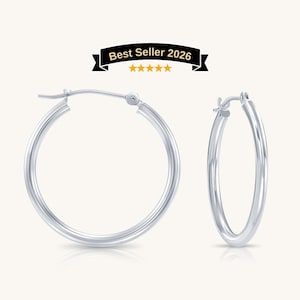 May include: A pair of polished silver-tone hoop earrings with a smooth, rounded profile and a hinged post closure. Above the earrings, a black banner displays the text Best Seller 2026 with five yellow stars centered underneath the text.