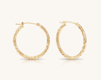 May include: A pair of gold hoop earrings featuring a textured, diamond-cut pattern along the outer surface. The circular earrings have a polished finish and include a hinged post mechanism for secure closure, reflecting light across the metal surface.