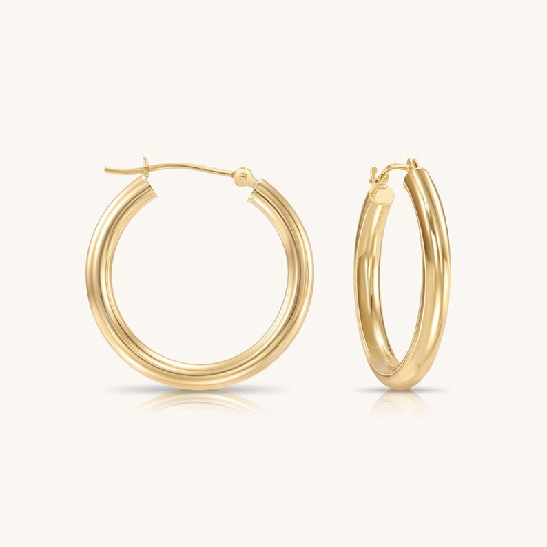 May include: A pair of polished gold hoop earrings with a smooth, rounded tube design. Each earring features a secure hinged post closure mechanism at the top, showcasing a classic circular silhouette with a reflective, high-shine metallic finish.