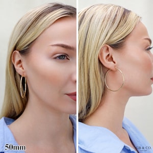 May include: A pair of gold hoop earrings with a thin, circular wire design. The earrings measure 50mm in diameter. The image displays the earrings from two different angles, showing the size and fit of the gold hoops on the earlobe.